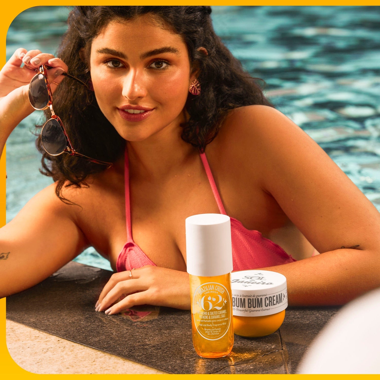 Woman holding sunglasses at the side of a swimming pool. Brazilian Crush cheirosa 62 perfume mist and brazilian bum bum cream in front of her.